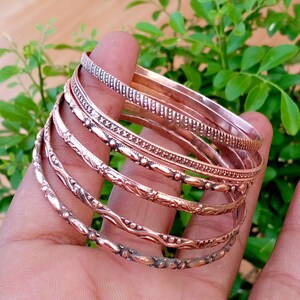 May include: A set of five copper-toned metal bangles featuring varied textures, including a ribbed band, a beaded band, and three bands with embossed floral and vine-like patterns, all stacked together in a circular arrangement.