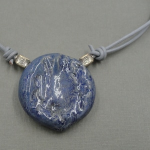 Pode incluir: Colar artesanal com um pendente azul grande de formato irregular. O pendente tem uma superfície texturizada e é suspenso por um cordão de couro cinza, conectado por contas de metal prateado com um design em espiral.