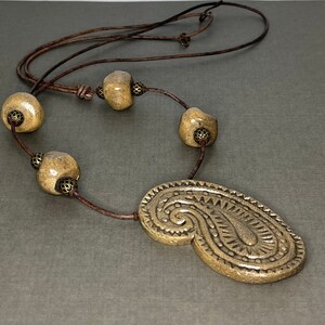 Pode incluir: Um colar de cordão de couro castanho com um grande pingente de cerâmica bege em forma de paisley. O pingente tem um design em relevo detalhado. O colar também apresenta quatro contas de cerâmica bege menores com detalhes dourados.