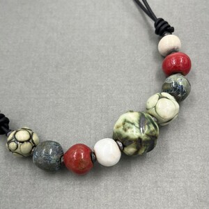 Pode incluir: Um colar com um cordão de couro preto e contas de cerâmica em tons de branco, verde, vermelho e azul. As contas têm um aspeto texturizado e pintado à mão.