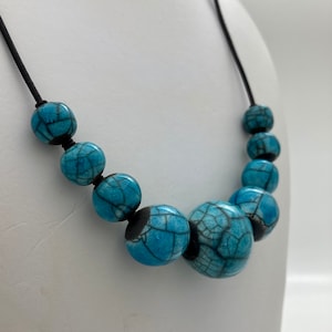 Pode incluir: Um colar azul turquesa com cordão preto e grandes contas redondas de cerâmica com um esmalte craquelado.