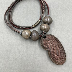 Pode incluir: Um colar de cordão de couro marrom com um pingente em forma de paisley. O pingente é feito de metal marrom escuro com um design em relevo. O colar tem quatro contas de cerâmica marrom e cinza.