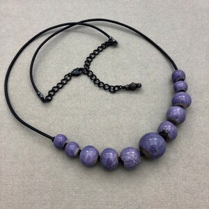 Pode incluir: Um colar de contas de cerâmica roxa com um cordão preto e um fecho de metal preto. As contas têm um acabamento de esmalte craquelado.