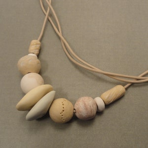 May include: A beige necklace with a variety of beige and cream colored ceramic beads. The beads are different shapes and sizes, including round, oval, and rectangular.