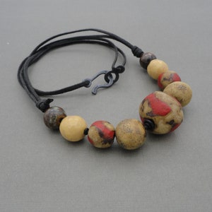 Op de afbeelding: Een ketting met een zwart koord en grote, ronde, veelkleurige keramische kralen. De kralen zijn een mix van bruin, beige en rood.