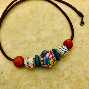 Op de afbeelding: Een ketting met een bruin leren koord en vijf kleurrijke keramische kralen. De kralen zijn een mix van wit, blauw, rood en groen.