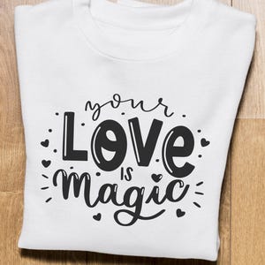 May include: White sweatshirt with the text "your Love is Magic" in black script. The design includes hearts and decorative elements. The sweatshirt is folded and placed on a wooden surface.