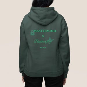 Puede incluir: Sudadera con capucha verde oscuro con el texto "MASTERMIND & Butterfly" en letras verdes y un gráfico de mariposa. La sudadera con capucha también tiene el texto "EST. 2023". La sudadera con capucha está hecha de un material suave y tiene una capucha.