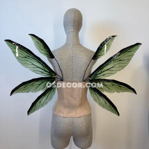 May include: Green and black fairy wings with a detailed vein design are attached to a nude-colored corset. The wings are displayed on a mannequin. The website address OSDECOR.COM is visible on the corset.