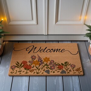 May include: A rectangular coir doormat with the word "Welcome" in black script. The bottom of the mat features a floral design with orange, purple, and yellow flowers. The mat is placed in front of a white door.