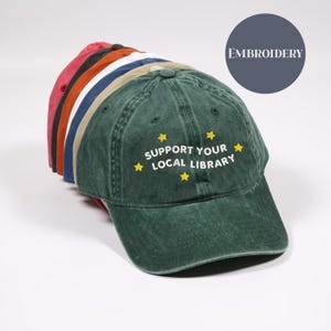 May include: A stack of baseball caps in various colors, including red, orange, navy, and green. The front-most green cap features the embroidered text "SUPPORT YOUR LOCAL LIBRARY" with yellow stars. The word "EMBROIDERY" is in a gray circle.