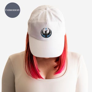 May include: White baseball cap with a gray brim featuring an embroidered emblem of a bird with a star above it. The word "EMBROIDERY" is printed on a gray circle in the upper left corner. The cap is worn by a person with red hair.