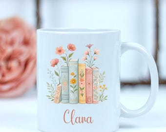 May include: White ceramic mug featuring a floral and book design. The design includes a stack of books with flowers and the name "Clara" in a script font. The mug has a curved handle.