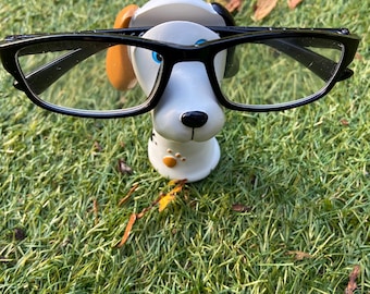 Puede incluir: Una figura de perro de cerámica con gafas de montura negra. El perro tiene un cuerpo blanco, manchas negras y orejas marrones. La figura está sobre hierba verde.