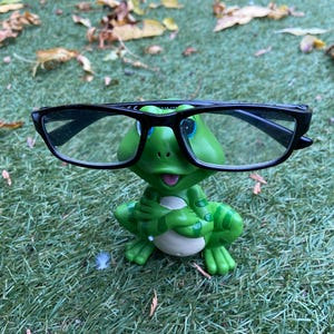 May include: A green frog ornament with black-framed spectacles. The frog is positioned on green grass, with some fallen leaves visible. The frog has a smiling expression and is holding its hands together.