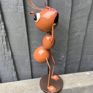 Puede incluir: Una caprichosa escultura de metal con forma de hormiga naranja. La escultura presenta una cabeza redonda con ojos grandes, un cuerpo segmentado y patas delgadas. Se apoya sobre una base circular, con las antenas de la hormiga curvadas hacia arriba. La escultura está pintada en tonos naranja y marrón.