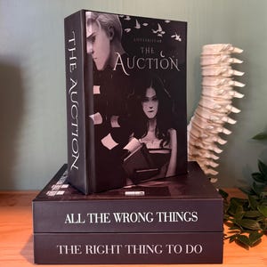 May include: Stacked books with dark covers. The top book is titled "The Auction" with illustrations of two people. Below are books titled "All The Wrong Things" and "The Right Thing To Do". The books are on a wooden surface.