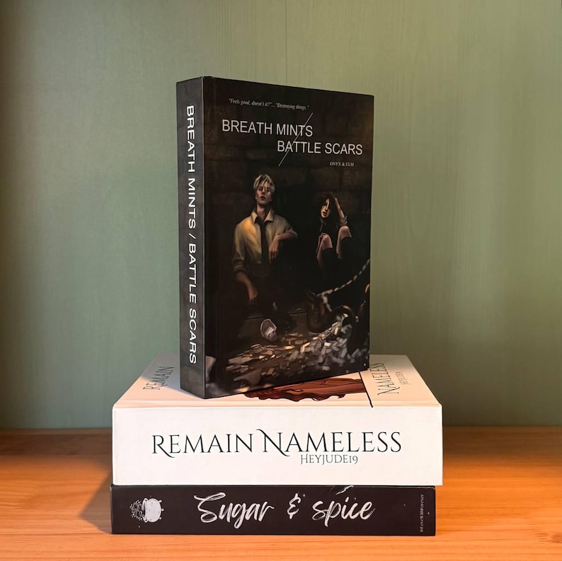 May include: Three books stacked on a wooden surface. The top book is titled "Breath Mints Battle Scars" with a dark cover featuring two figures. Below it is a white book with "Remain Nameless" and "Heyjude19". The bottom book is black with "Sugar & Spice".