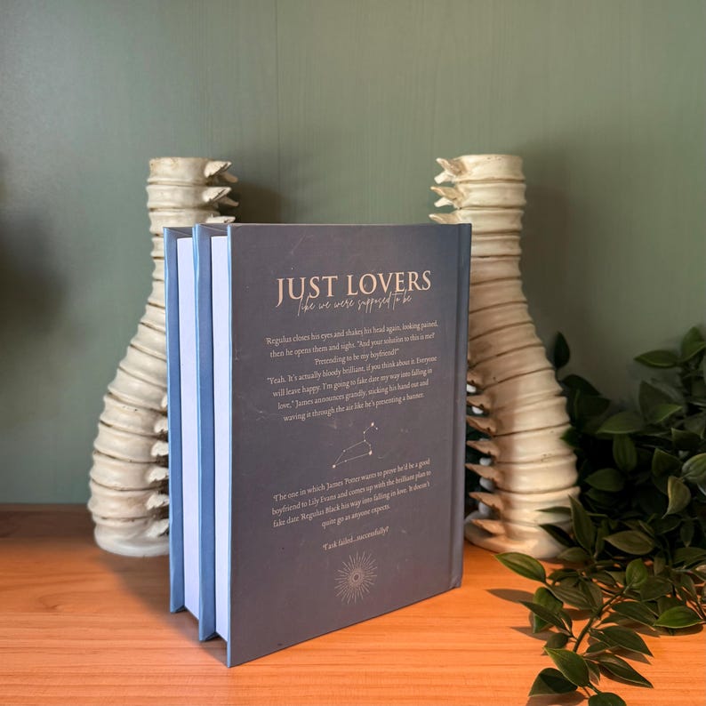 May include: A hardcover book titled "JUST LOVERS like we were supposed to be" with a dark blue cover and white text. The book is propped up on a wooden surface, with decorative vases in the background.