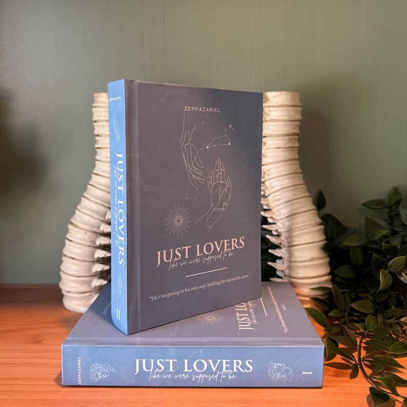May include: Two books titled "JUST LOVERS" are stacked on a wooden surface. The top book is upright, with a blue spine and a dark gray cover featuring hand illustrations and text. The bottom book is lying flat, also with the same title and design.