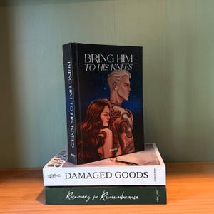 May include: A stack of three books on a wooden surface. The top book, "Bring Him to His Knees," features a dark cover with a couple illustration. Below it is "Damaged Goods" and "Rosemary for Remembrance."