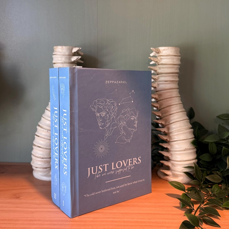 May include: A stack of books titled "JUST LOVERS" with a blue cover and white text. The cover features line art of two faces and the author's name, ZEPPAZARIEL. The books are on a wooden surface with decorative vases in the background.