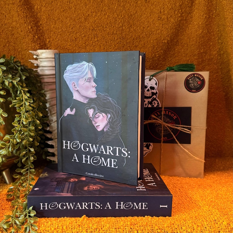 May include: Two hardcover books titled "Hogwarts: A Home" are stacked on a textured orange surface. The top book features a couple embracing under a starry sky. A wrapped package with a skull sticker and a green tassel is in the background.