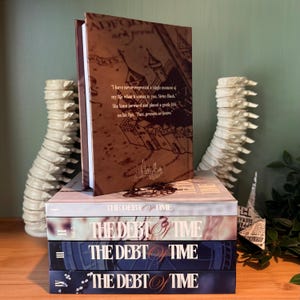 K&ouml;nnte beinhalten: Ein Stapel von vier B&uuml;chern mit dem Titel "THE DEBT OF TIME". Das oberste Buch ist ge&ouml;ffnet und zeigt Text und eine kartenartige Illustration. Die B&uuml;cher sind auf einer Holzoberfl&auml;che gestapelt, mit dekorativen Objekten auf beiden Seiten.