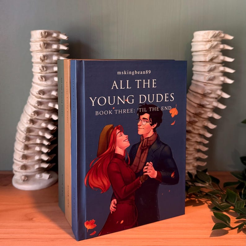 以下が含まれることがあります： 「ALL THE YOUNG DUDES」というタイトルと「BOOK THREE: 'TIL THE END」というサブタイトルが書かれた、濃い青色の表紙の本の束。表紙には踊るカップルが描かれています。本の背表紙には、著者の名前「mskingbean89」が見えます。