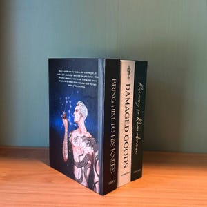May include: Three hardcover books on a wooden surface. The first book has a dark cover with a man with tattoos and the title "BRING HIM TO HIS KNEES". The second book is black with the title "DAMAGED GOODS". The third book is green with the title "Rosemary for Remembrance".