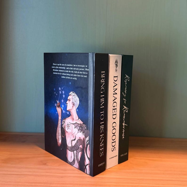 May include: Three hardcover books on a wooden surface. The first book has a dark cover with a man with tattoos and the title "BRING HIM TO HIS KNEES". The second book is black with the title "DAMAGED GOODS". The third book is green with the title "Rosemary for Remembrance".