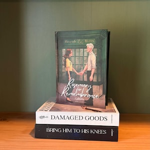 May include: A stack of three books on a wooden surface. The top book is titled "Rosemary for Remembrance" with an illustration of a couple. Below it is "DAMAGED GOODS" and the bottom book is "BRING HIM TO HIS KNEES".