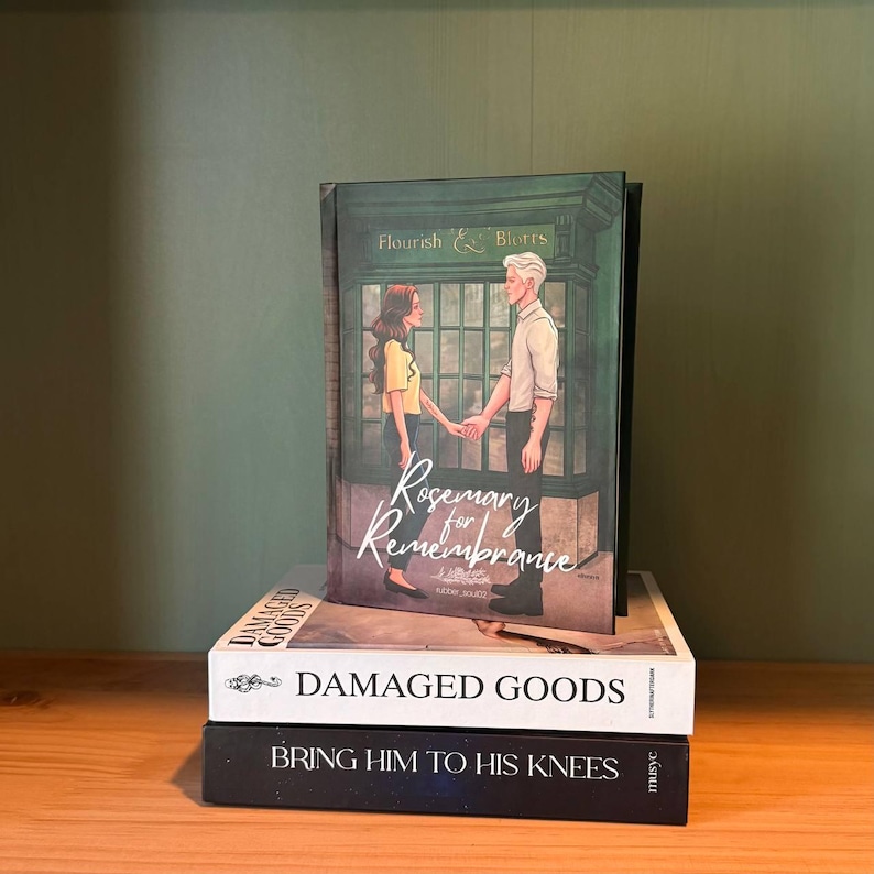 May include: A stack of three books on a wooden surface. The top book is titled "Rosemary for Remembrance" with an illustration of a couple. Below it is "DAMAGED GOODS" and the bottom book is "BRING HIM TO HIS KNEES".