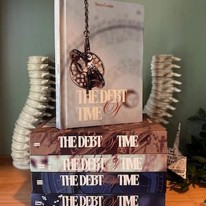 K&ouml;nnte beinhalten: Eine Reihe von B&uuml;chern mit dem Titel "THE DEBT OF TIME" von Shaya Lonnie. Das oberste Buch zeigt eine detaillierte Illustration eines Zeitmessger&auml;ts. Die B&uuml;cher sind in verschiedenen Braun- und Blaut&ouml;nen gehalten, mit dem Titel in eleganter Schrift.