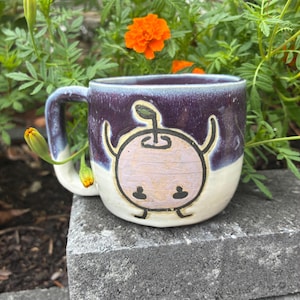 May include: A ceramic mug with a two-tone glaze featuring a dark purple upper section and a white lower section. The front displays a hand-drawn illustration of a light pink, round character with small arms, legs, and a stem, outlined in black.
