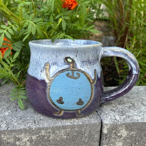 May include: A ceramic mug with a two-tone glaze featuring a light blue top and a dark purple bottom. The front displays a hand-painted light blue circular character with thin brown limbs, a small head, and a dark brown symbol on its chest.