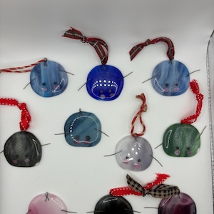 May include: A collection of handmade, round ornaments in various colors, including blue, green, pink, and black. Each ornament features a painted face with small black eyes and a smiling mouth. They are adorned with red and plaid ribbons for hanging.