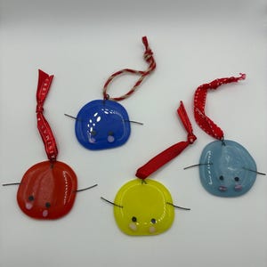 May include: Four colorful, round Christmas ornaments with painted faces and wire arms. The ornaments are red, blue, yellow, and light blue. Each ornament has a ribbon or twine hanger. One red ornament has "MERRY CHRISTMAS" printed on the ribbon.