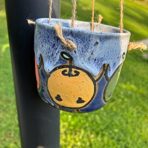 Op de afbeelding: Een hangende keramische plantenpot met een blauwe glazuurafwerking en een gespikkelde textuur. De buitenkant is voorzien van handgeschilderde oranje en groene fruitmotieven met zwarte omlijningen. De pot hangt aan drie strengen natuurlijk jutekoord.