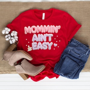 May include: A red t-shirt with the phrase "Mommin' Ain't Easy" in a fluffy, textured font. The shirt is paired with blue jeans and beige ankle boots. A sprig of cotton adds a decorative touch.
