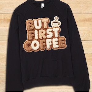 May include: Black sweatshirt with the phrase "BUT FIRST COFFEE" in a textured, brown and cream-colored design. A small coffee cup graphic is next to the text. The sweatshirt has a front pocket and long sleeves.
