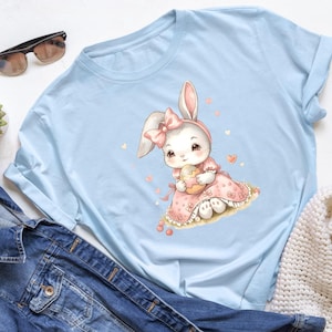 Könnte beinhalten: Hellblaues T-Shirt mit einer Aquarellillustration eines niedlichen Hasen, der ein rosa Kleid trägt und ein verziertes Osterei hält. Der Hase hat eine rosa Schleife und ist von kleinen Herzen umgeben. Das Shirt ist aus weichem Material.