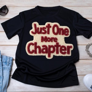 May include: A navy blue t-shirt with the text "Just One More Chapter" in a cream-colored, textured design with red lettering. The shirt is displayed on a light wood-grain surface.