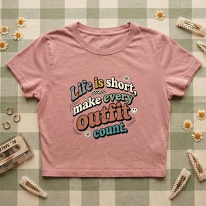 May include: A dusty rose-colored t-shirt with the text "Life is short, make every Outfit count." in a retro font. The shirt is styled with accessories like pearl hair clips, daisy flowers, and a vintage cassette tape.