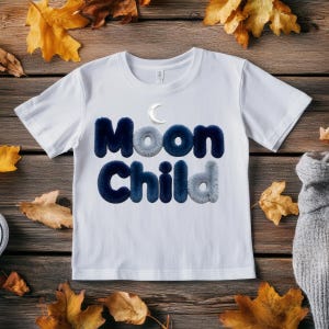 May include: White t-shirt with the words "Moon Child" in a fluffy, textured font, with a crescent moon above the word "Moon". The shirt is surrounded by autumn leaves on a wooden surface.