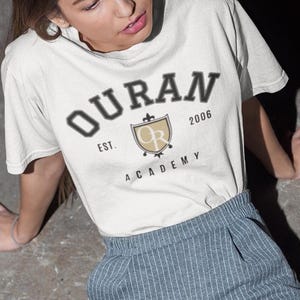May include: A cream-coloured short-sleeve t-shirt featuring the dark text OURAN ACADEMY, EST. 2006, and a shield emblem with the letters OR. The shirt is paired with grey pinstriped trousers and white trainers, creating a coordinated casual outfit.