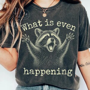 Puede incluir: Una camiseta de manga corta gris carbón con una ilustración gráfica de un mapache con las patas levantadas. El texto "What is even happening" está impreso en un color crema claro encima y debajo de la ilustración del mapache.