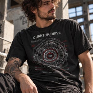 Peut inclure: Un t-shirt gris anthracite à manches courtes avec un graphique technique blanc et rouge sur la poitrine. L'impression inclut le texte "QUANTUM DRIVE", "FUSION COPE" et des caractères cyrilliques, représentant un design de machine circulaire.