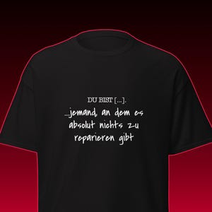 Peut inclure: Un t-shirt noir à manches courtes avec le texte blanc suivant : DU BIST [...]. ...jemand, an dem es absolut nichts zu reparieren gibt. Le vêtement présente une encolure ronde simple et une coupe standard, offrant un design visuel épuré.