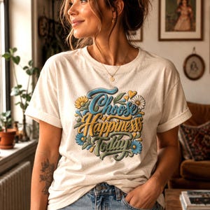 Könnte beinhalten: Ein cremefarbenes Kurzarm-T-Shirt mit einem Grafikdruck, der den Text "Choose Happiness Today" in blauer und gelber Schreibschrift zeigt, umgeben von Illustrationen aus blauen, gelben und weißen Blumen sowie grünen Blättern.